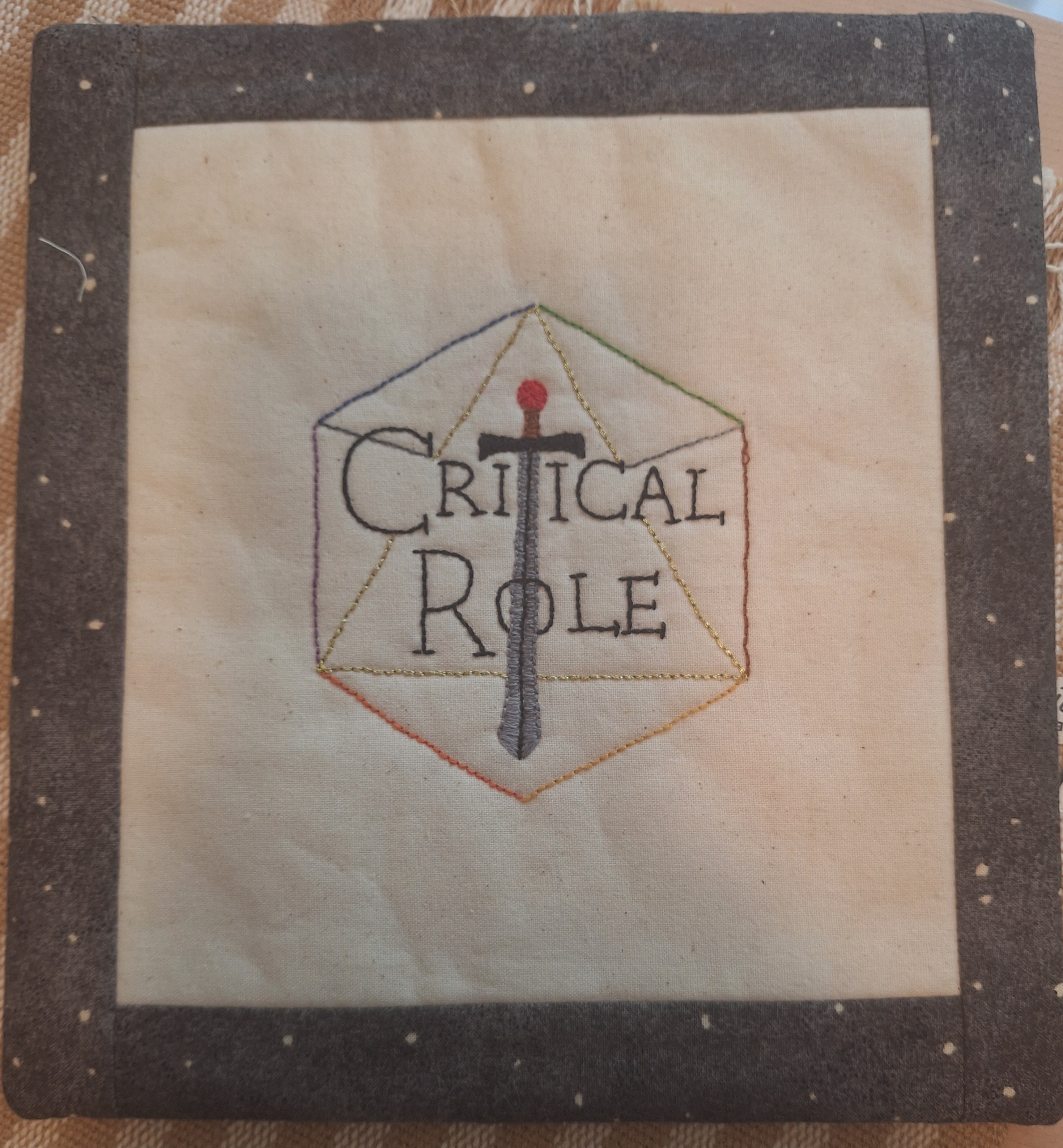 embroidery of a 20-sided die with the words Critical Role on it. The T is a sword.
