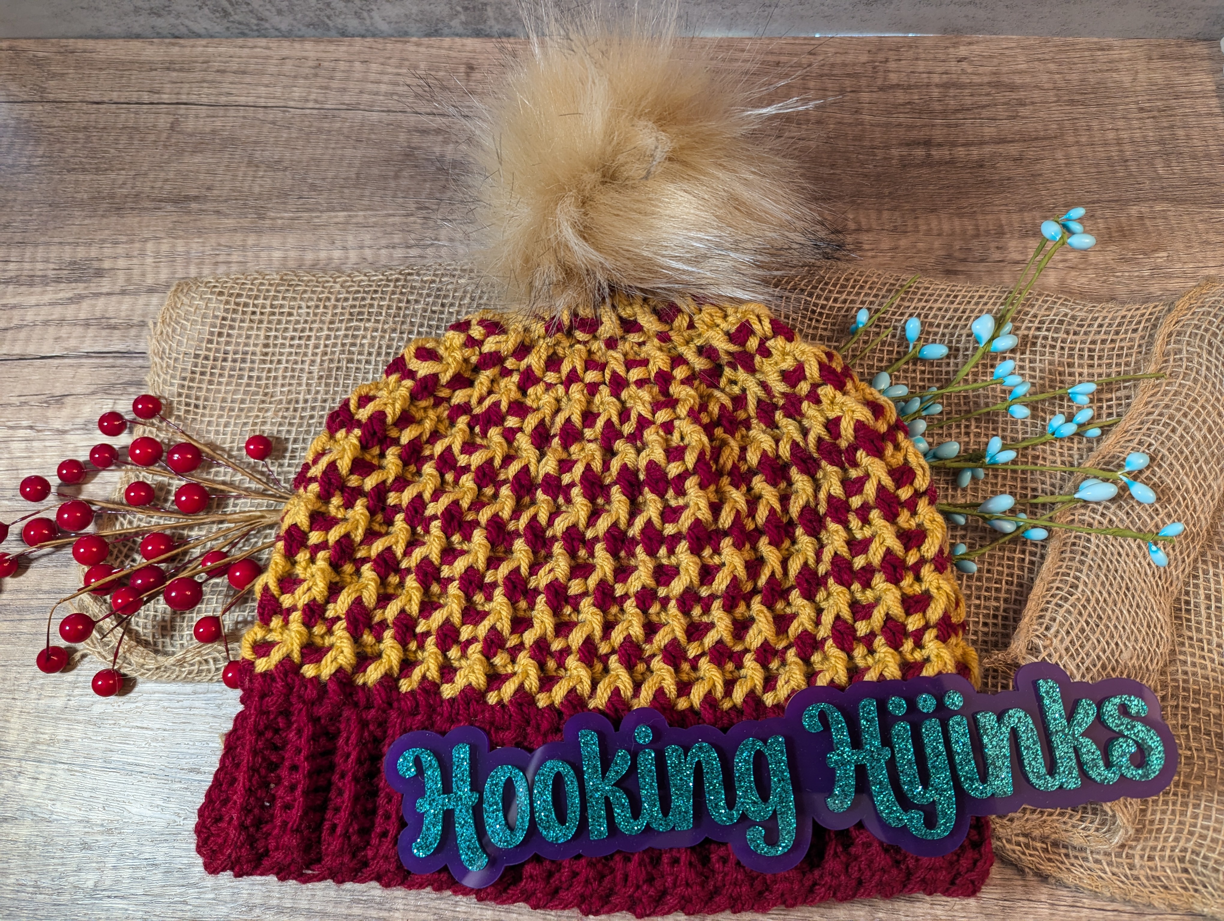 A red and yellow knit beanie with the words Hooking Hijinks in blue script in front of it.